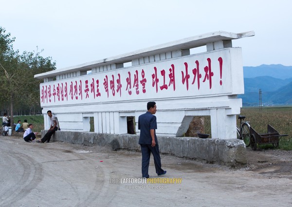 North Korean man passing in front of a propaganda billboard saying let's push revolution and construction as the spirit of the great leader in his lifetime, South Hamgyong Province, Hamhung, North Korea