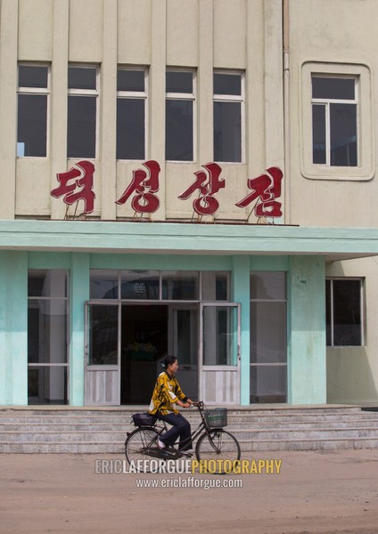 North Korean woman riding a bicycle in front of deoksong shop, South Hamgyong Province, Hamhung, North Korea